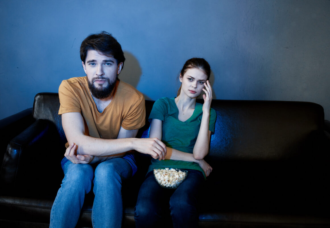 Twee bezorgde ouders zitten op de bank en kijken serieus naar televisie, met een kom popcorn op schoot.