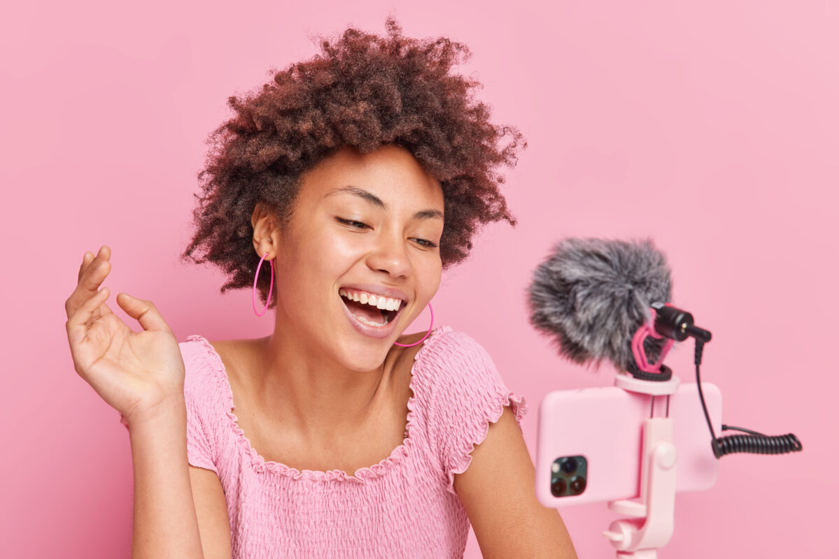 Vrolijke jonge vrouw maakt video met smartphone en microfoon tegen roze achtergrond, als influencer op social media.