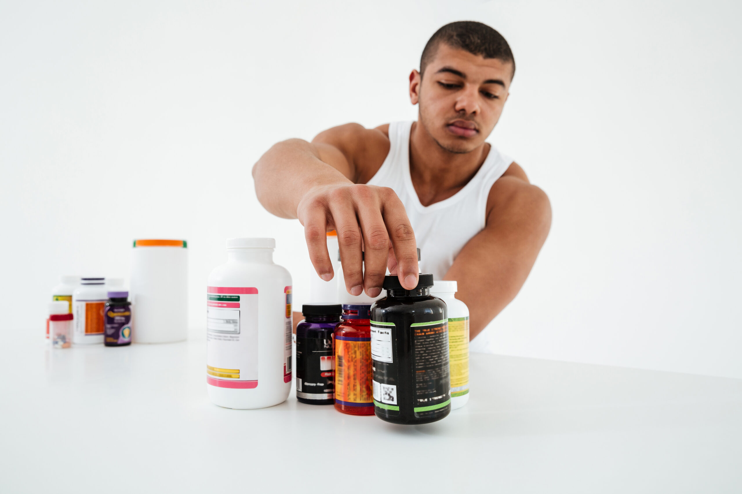Jonge sporter in tanktop die supplementen bekijkt op een witte tafel – focus op risico’s supplementen jongeren