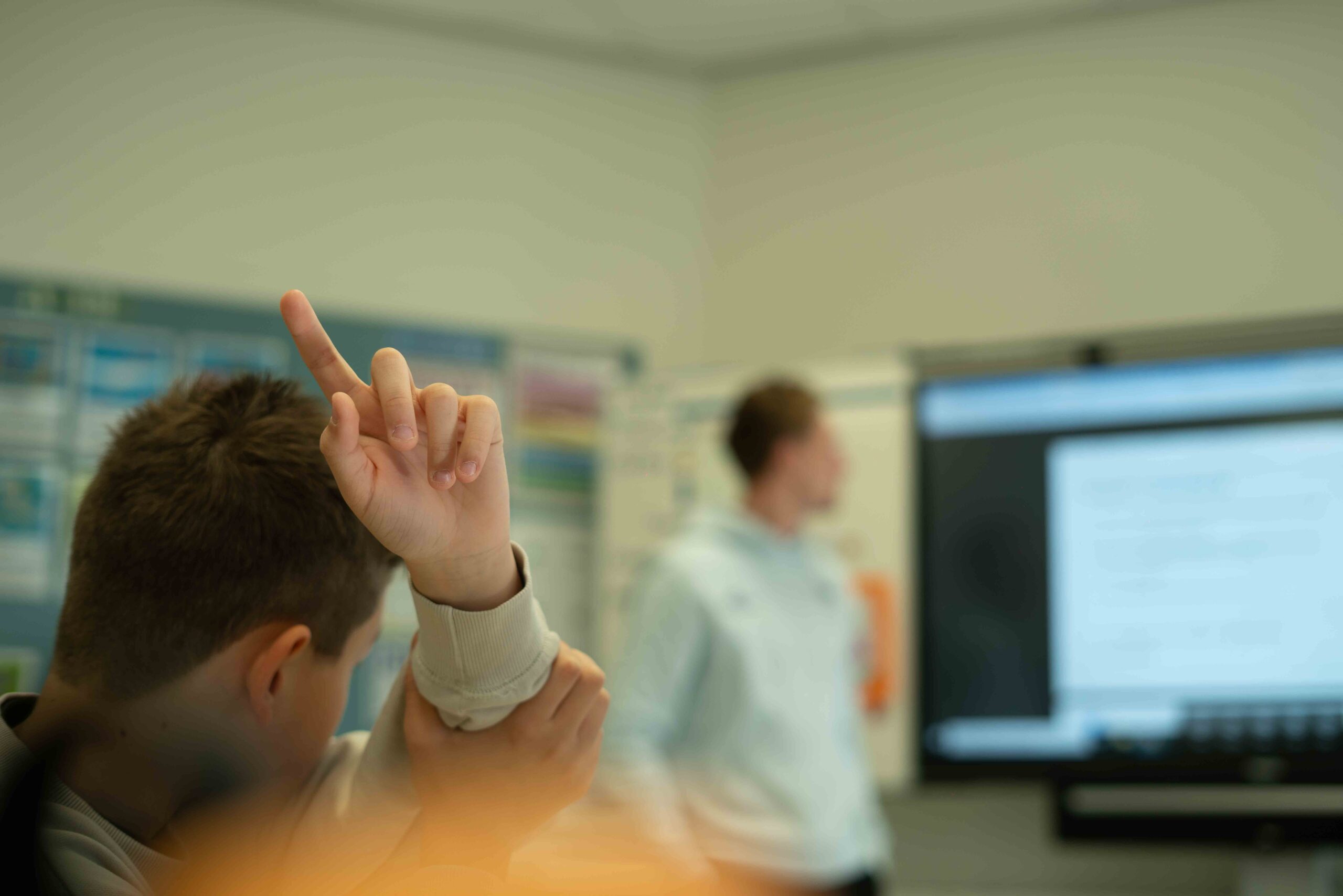 Leerling steekt hand op tijdens een interactieve voorlichting over cyberpesten, met een spreker voor de klas en digitaal lesmateriaal op het scherm.