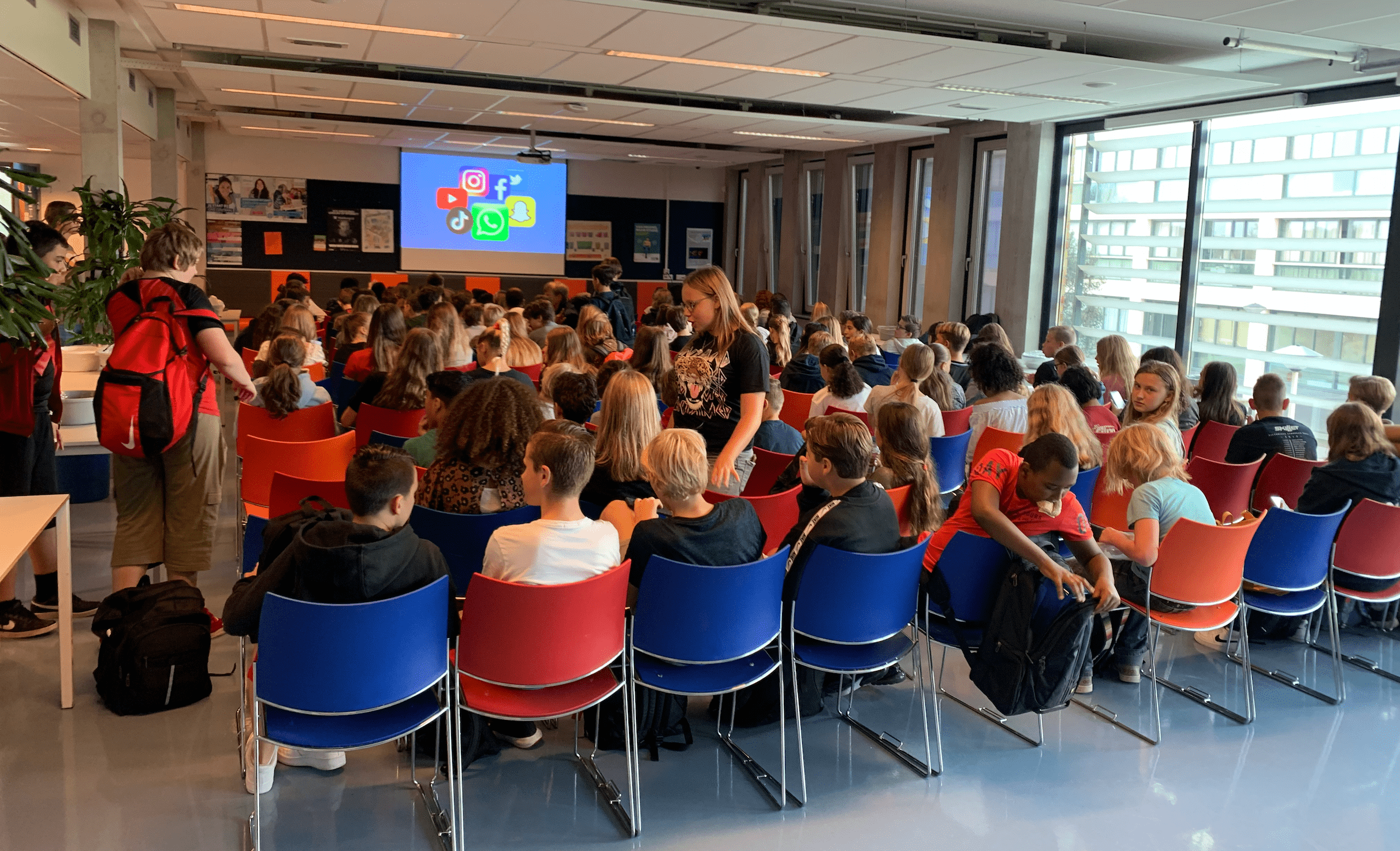 Groep middelbare scholieren tijdens een voorlichting over social media en online gedrag, met logo’s van populaire apps op het projectiescherm.