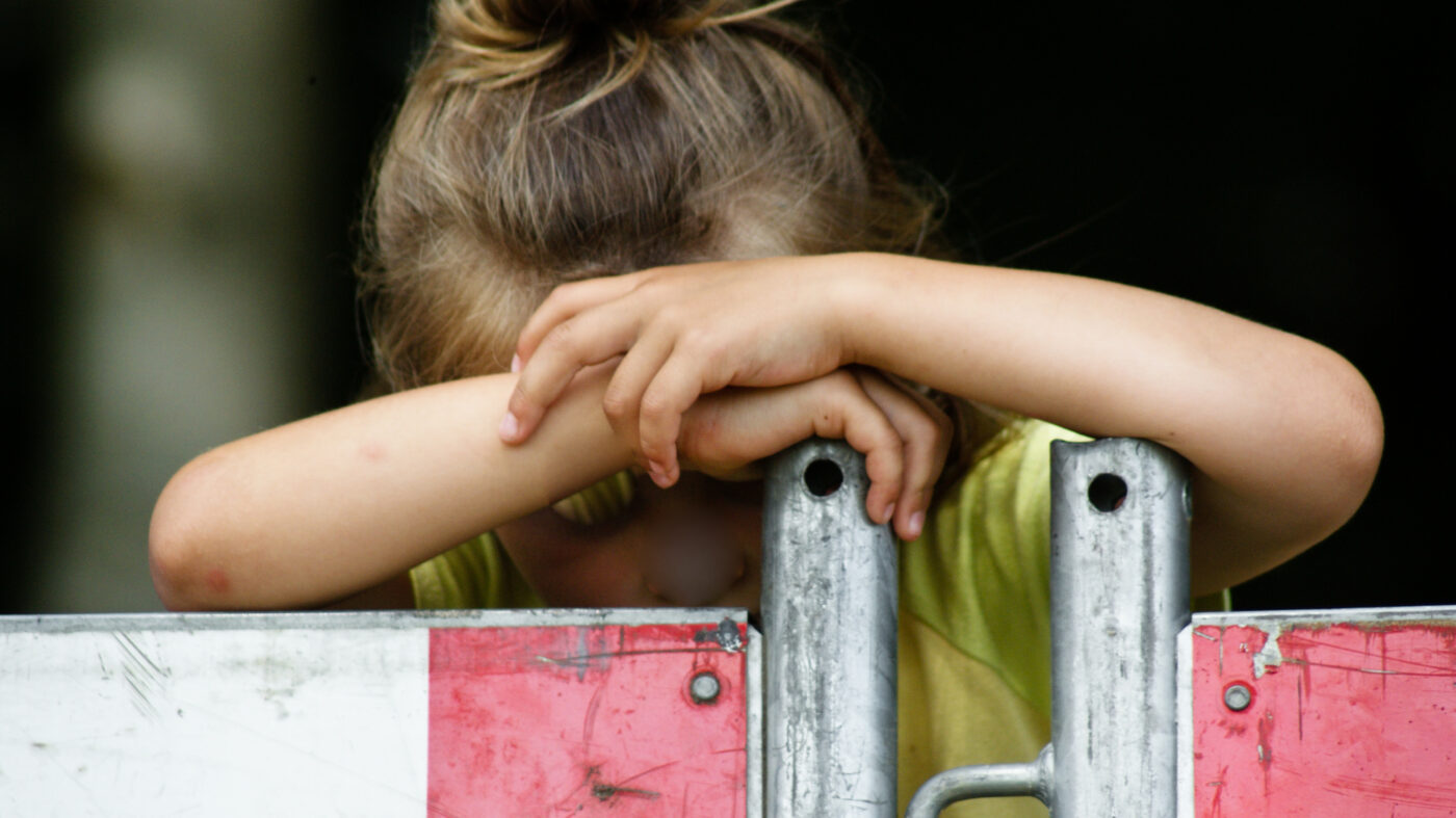 Verdrietig meisje leunt met hoofd op armen achter een hek – symbool voor kwetsbaarheid en schijnveiligheid bij seksueel misbruik jongeren.