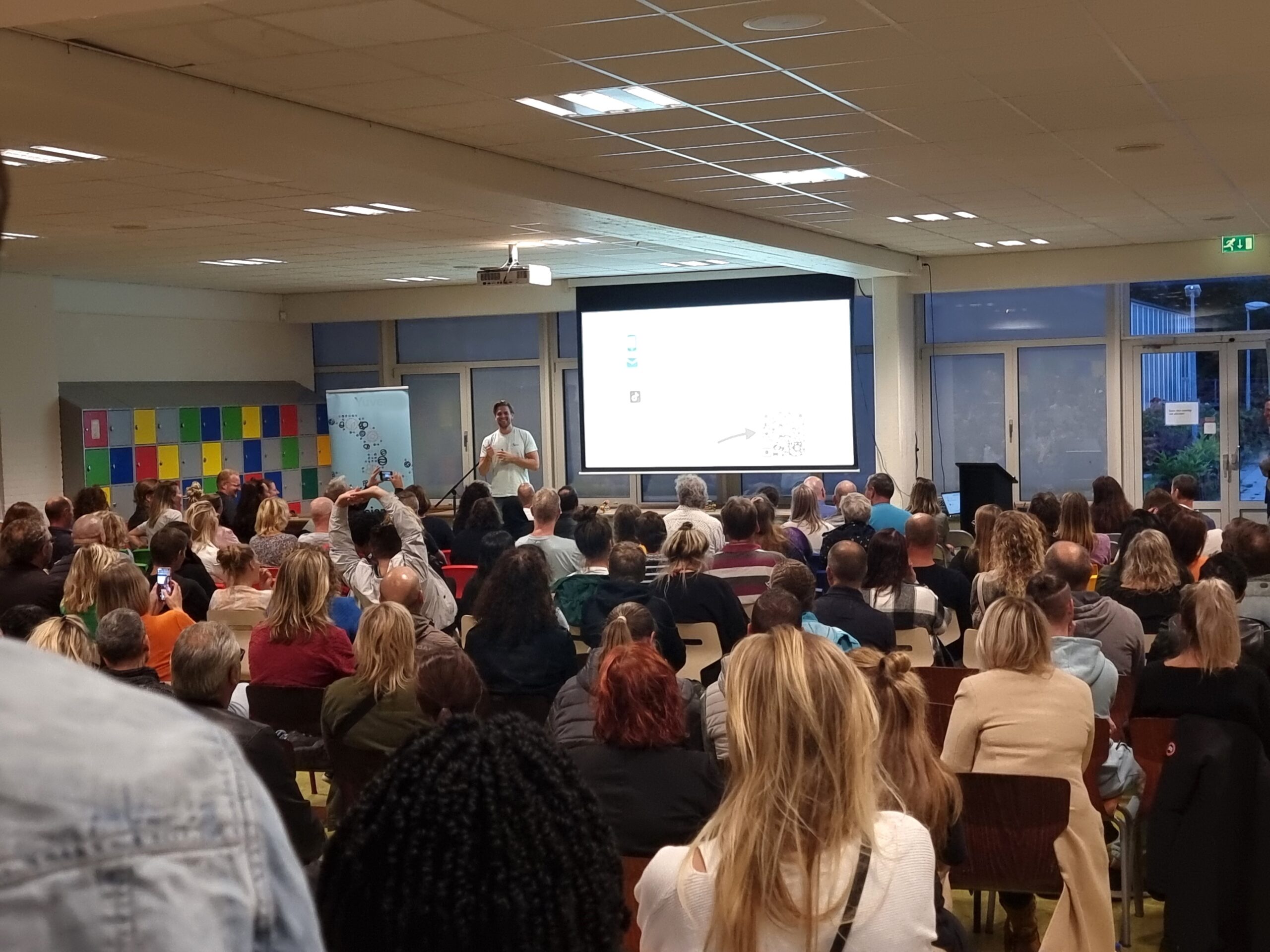 Spreker geeft voorlichting aan ouders over geld en de online wereld tijdens een ouderavond op school, met een volle zaal geïnteresseerde aanwezigen.
