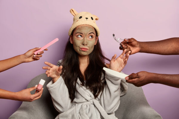 Tienermeisje met gezichtsmasker in badjas kijkt bezorgd terwijl handen haar beautyproducten aanreiken.