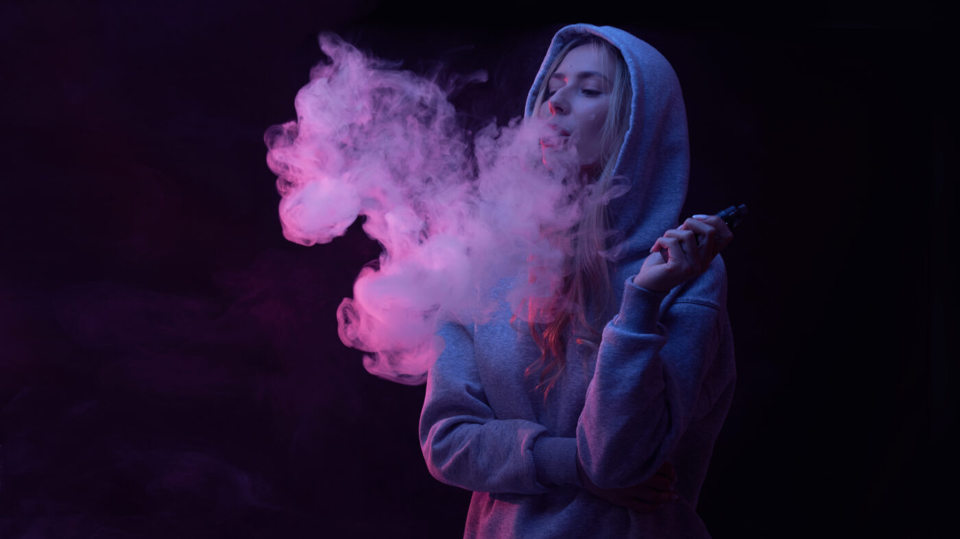 Tienermeisje in grijze hoodie blaast damp uit vape. Vapen onder jongeren neemt sterk toe