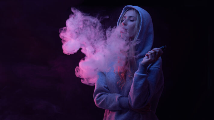 Tienermeisje in grijze hoodie blaast damp uit vape. Vapen onder jongeren neemt sterk toe