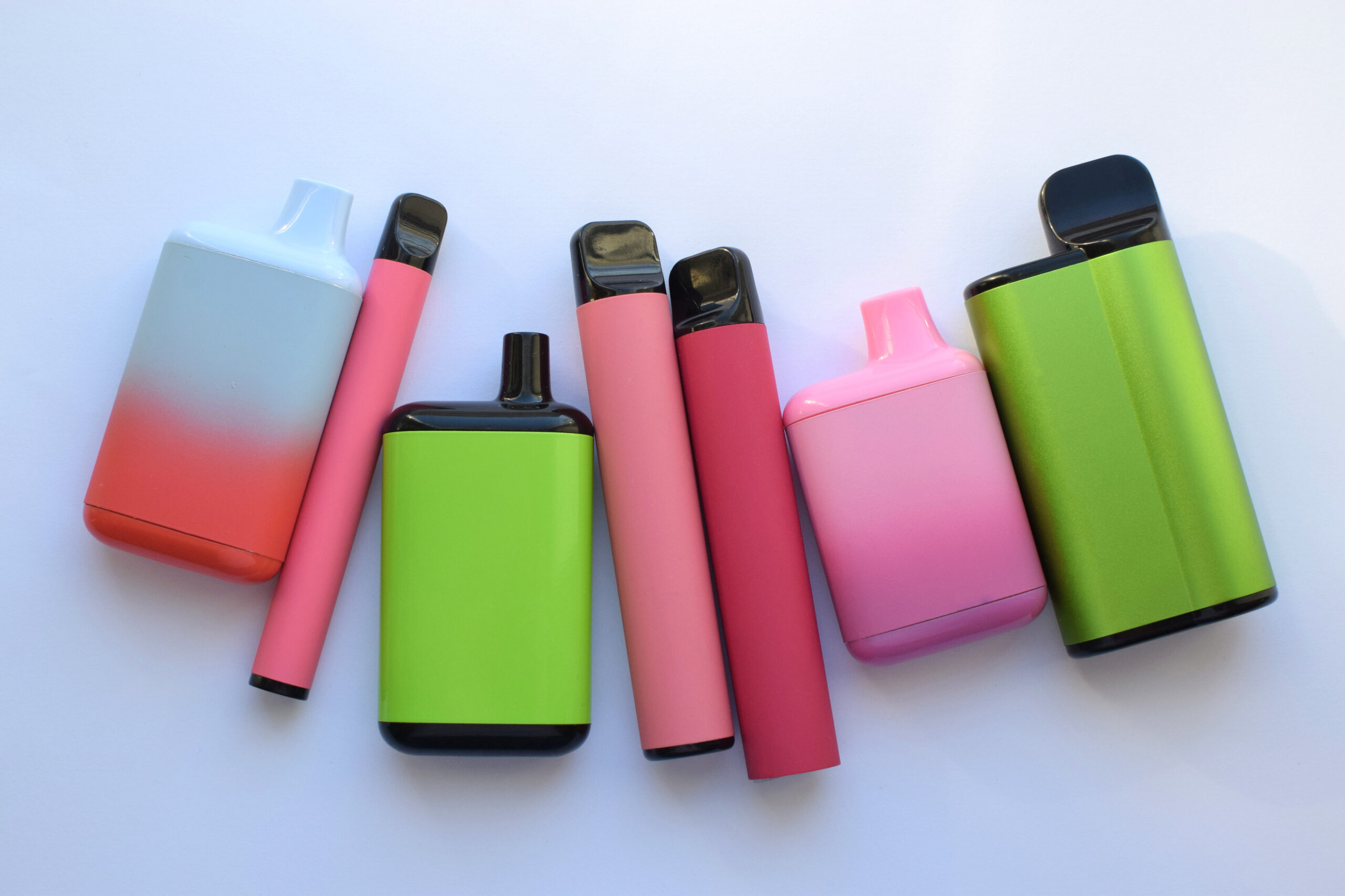 Zeven felgekleurde vapes in roze, groen en rood op een witte tafel – populair onder jongeren maar schadelijk voor de gezondheid.