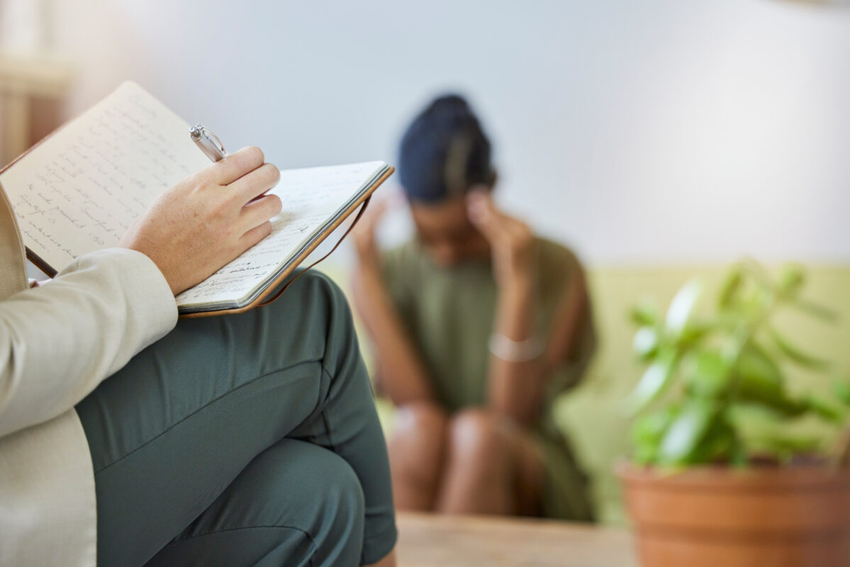 Psycholoog schrijft in notitieboek terwijl een verdrietige jongere op de achtergrond met het hoofd in de handen zit.