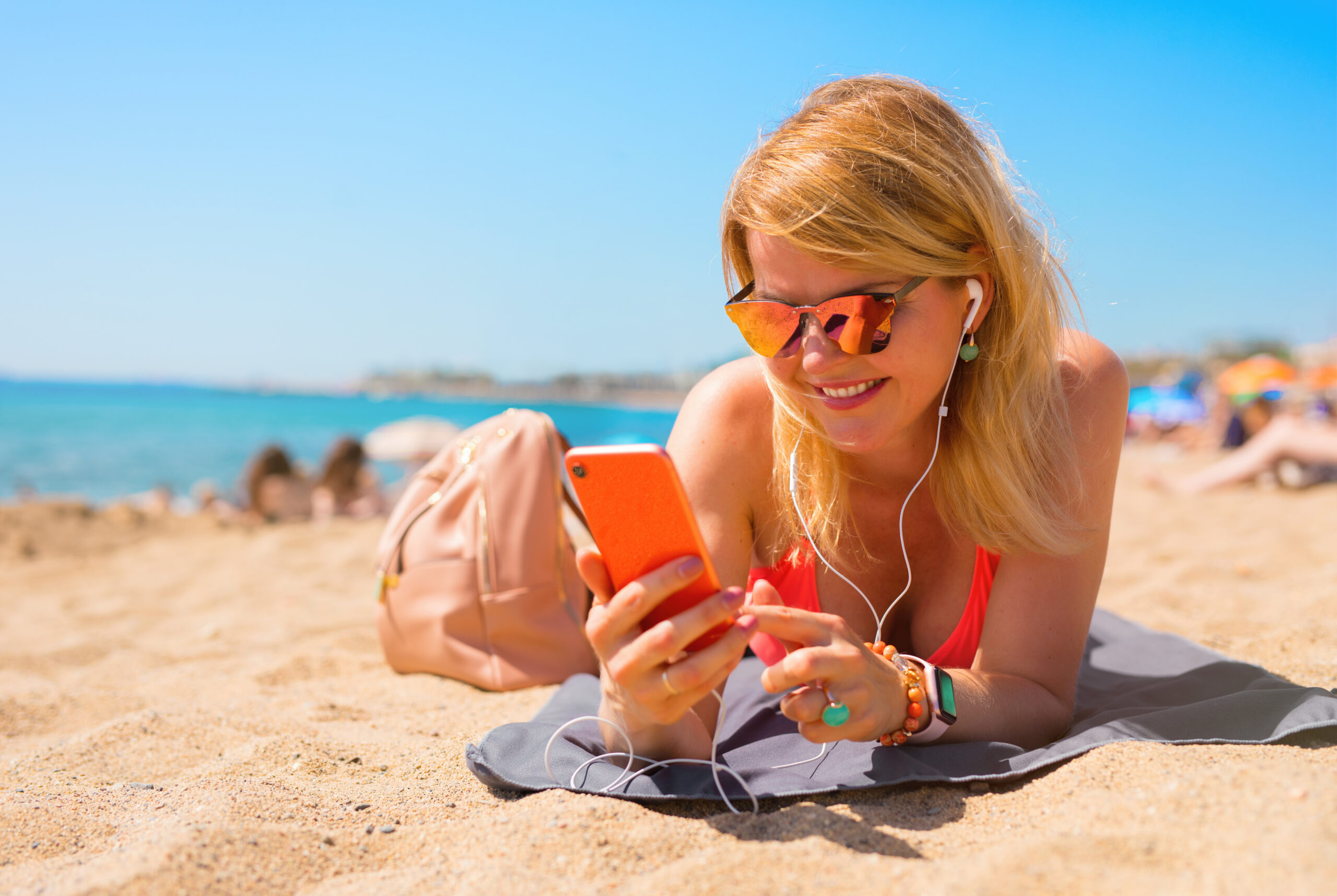 Vrouw op het strand met smartphone en oordopjes, symboliseert online actief zijn tijdens zomervakantie en het belang van online veiligheid.