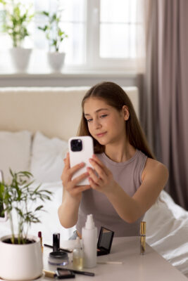 Tienermeisje maakt een selfie met smartphone terwijl ze make-up draagt aan haar kaptafel, beïnvloed door schoonheidsidealen op sociale media.