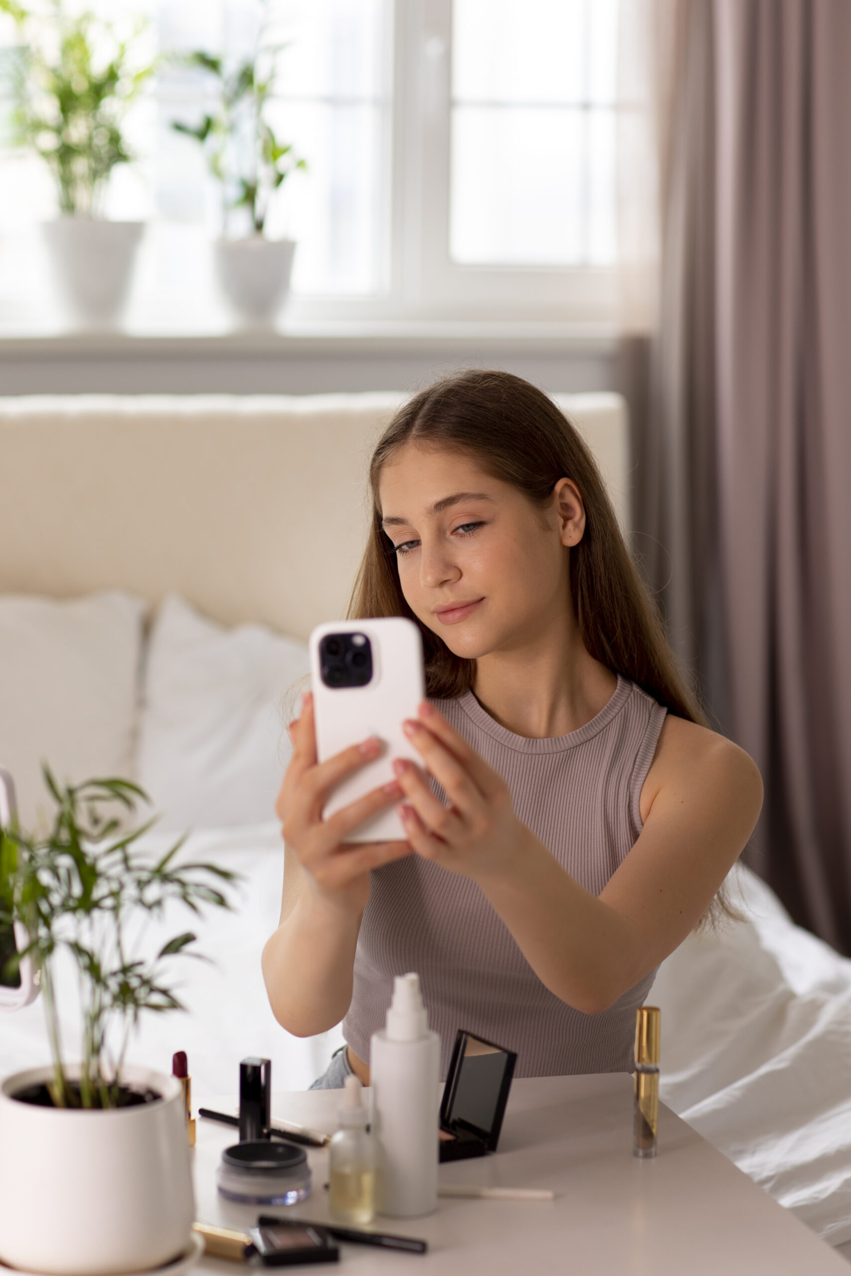 Tienermeisje maakt een selfie met smartphone terwijl ze make-up draagt aan haar kaptafel, beïnvloed door schoonheidsidealen op sociale media.