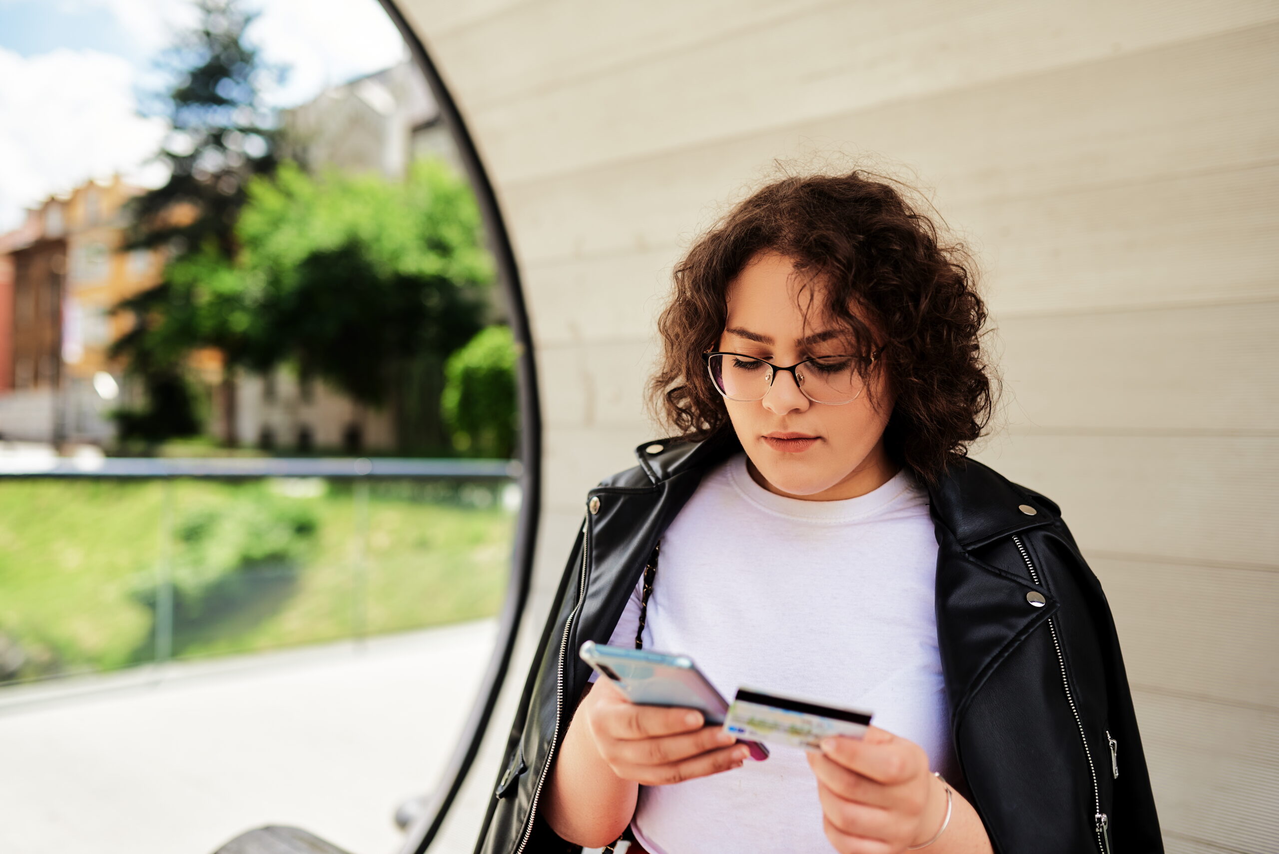 Jongere met bril en leren jas bekijkt haar bankpas terwijl ze een telefoon vasthoudt, mogelijk benaderd als geldezel.
