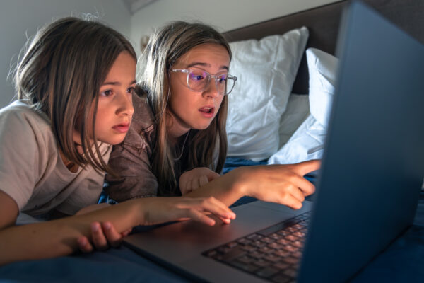 Twee kinderen liggen op bed en kijken geconcentreerd naar een laptop, terwijl één van hen iets aanwijst op het scherm.
