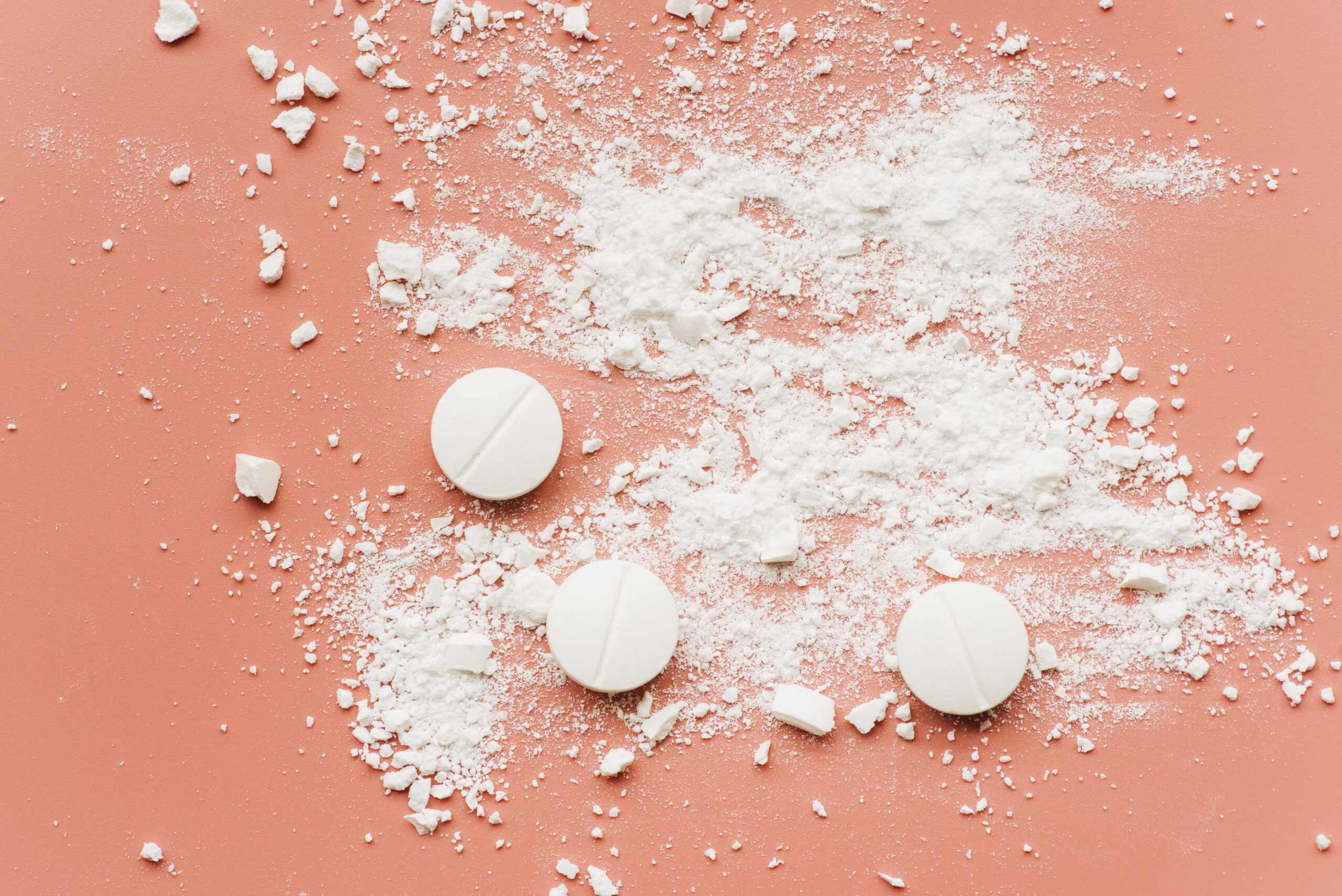 Verpulverde pillen en witte poederachtige substantie op een roze achtergrond, als symbool voor online verhandelde drugs zoals coke en ketamine.