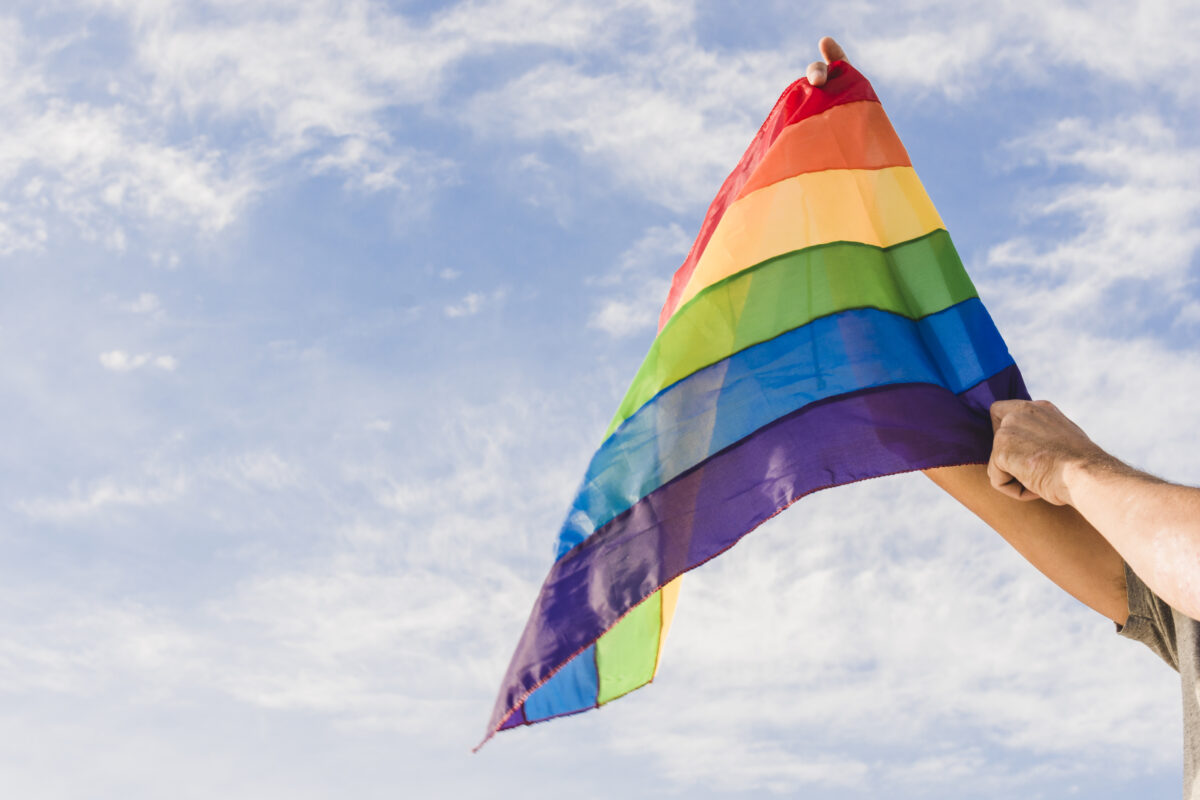 Regenboogvlag wordt omhooggehouden tegen een blauwe lucht als symbool voor LHBTI-zichtbaarheid en inclusie.