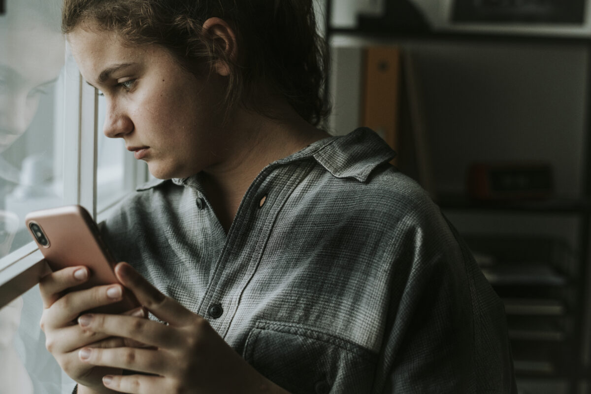 Jongere kijkt somber naar haar telefoon terwijl ze bij het raam staat, mogelijk worstelend met onzekerheid of negatieve gedachten.