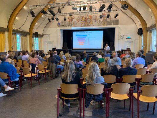 Ouderavond op school met ouders in een aula die luisteren naar een presentatie over digitale opvoeding en verslaving.
