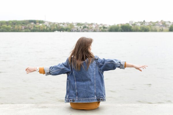 Jongere zit met gespreide armen aan het water en kijkt uit over een rustige omgeving.