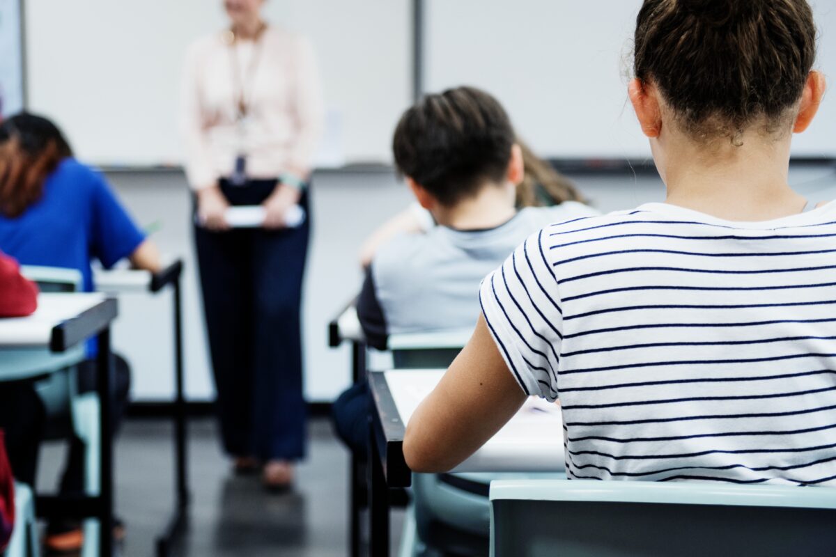 Leerlingen zitten aan hun tafels in een klaslokaal terwijl een docent voor de klas staat.