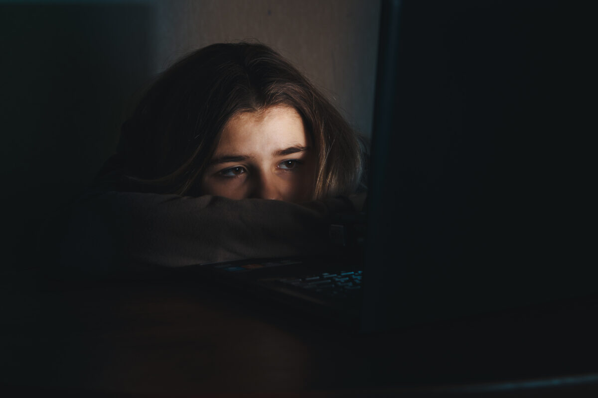 Meisje kijkt in het donker bezorgd naar een laptopscherm, verlicht door blauw licht van het scherm.