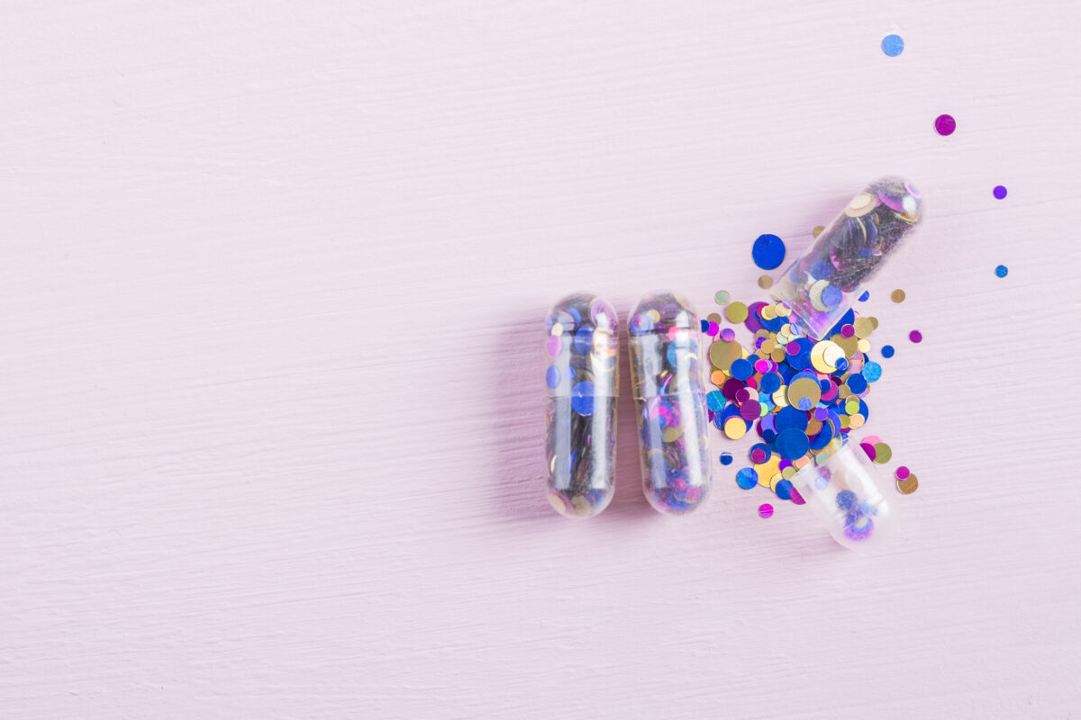 Transparante capsules gevuld met kleurrijke confetti liggen open op een lichtroze achtergrond, als visuele metafoor voor de aantrekkingskracht van drugs.