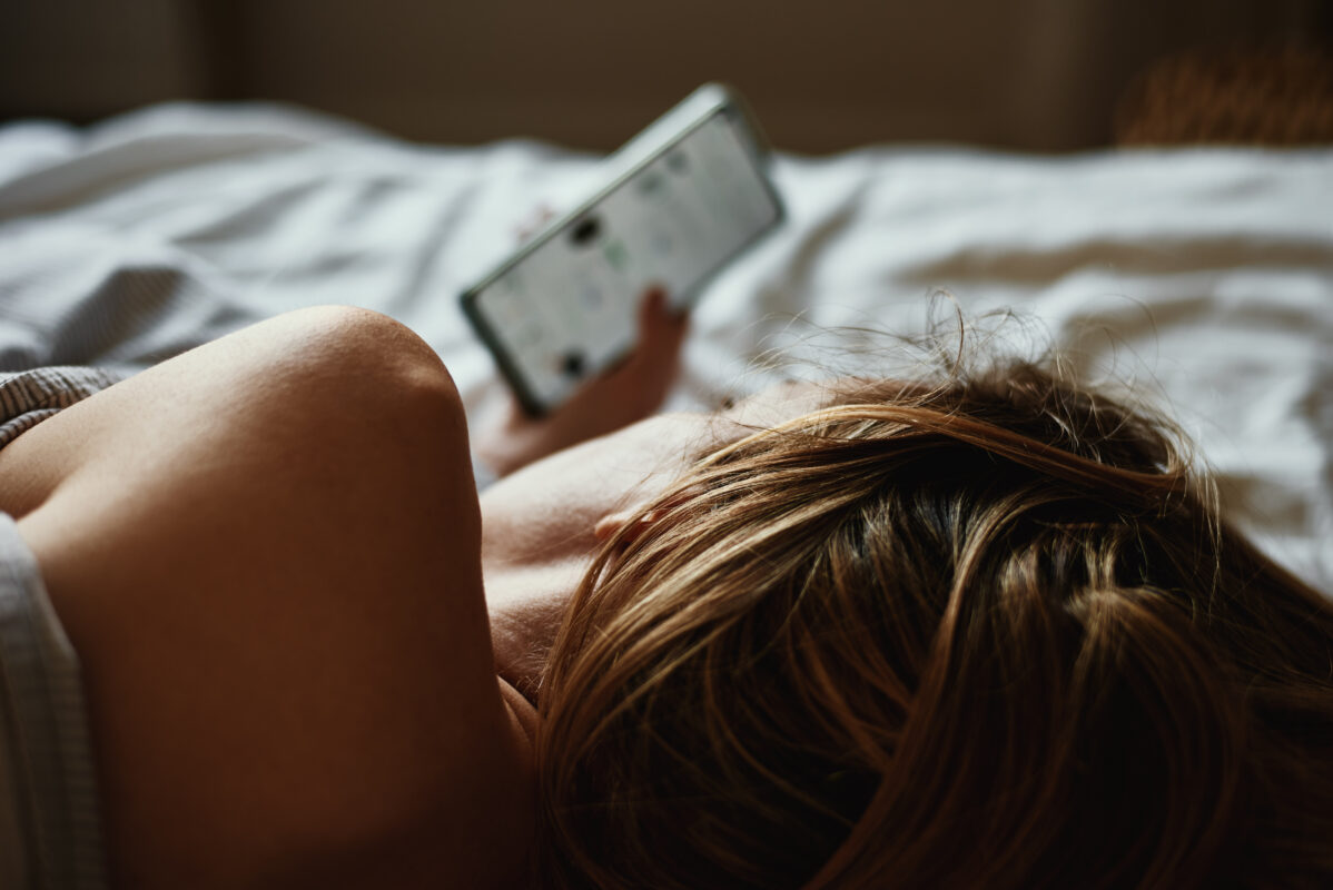 Jongere ligt op bed en kijkt op haar smartphone, symbool voor online risico’s zoals deepnudes en digitale grensoverschrijding.