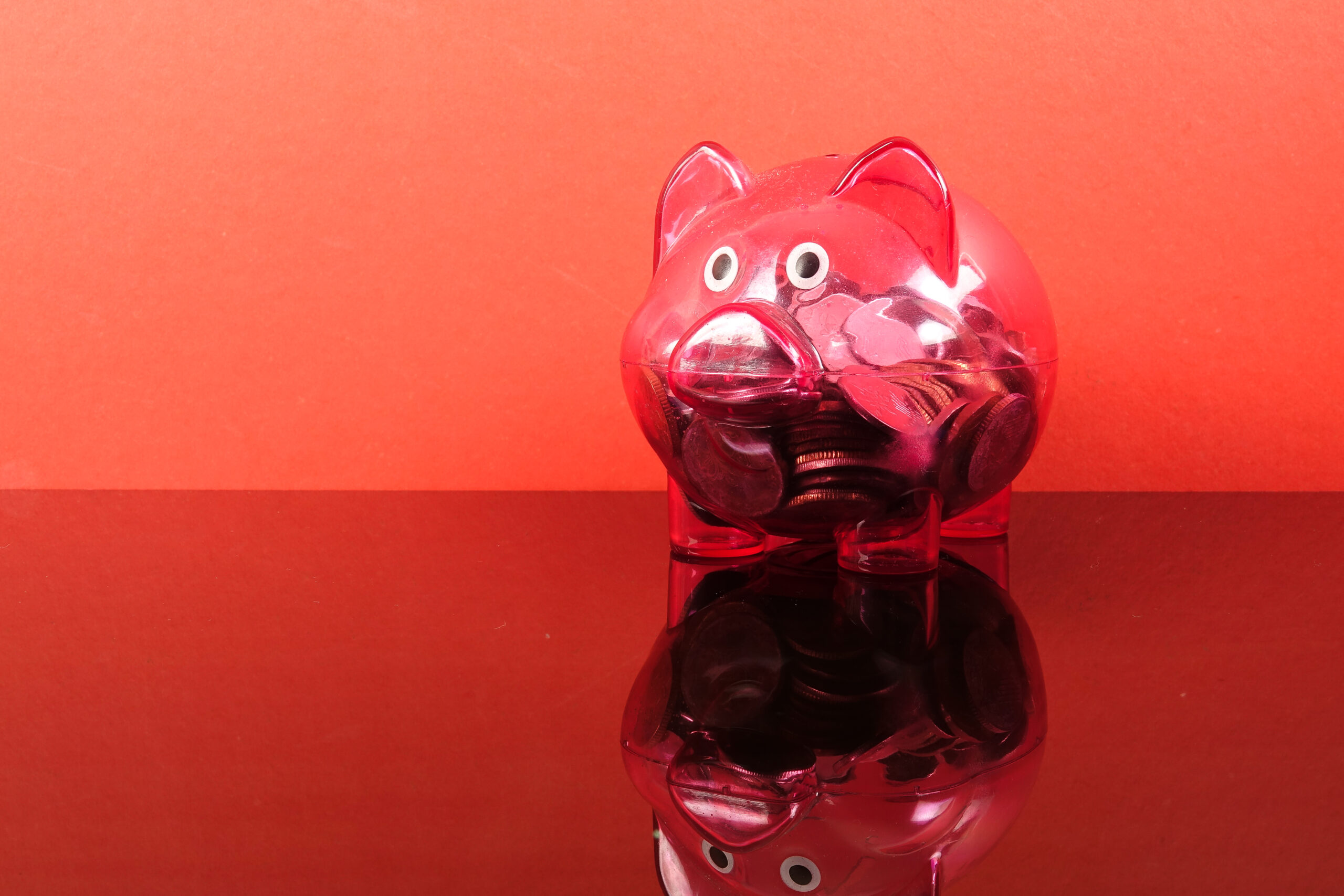 Transparante rode spaarpot met muntgeld op een rode achtergrond, symbool voor sparen en financiële keuzes bij jongeren