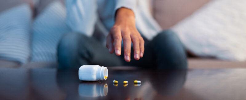 Persoon reikt naar een omgevallen potje met pijnstillers en capsules op tafel, symbool voor risico’s van opioïde pijnstillers en medicijnmisbruik.