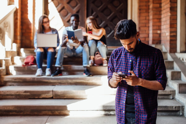 Jongere kijkt op zijn telefoon terwijl andere leerlingen op de achtergrond samen zitten, mogelijk buitengesloten of gepest