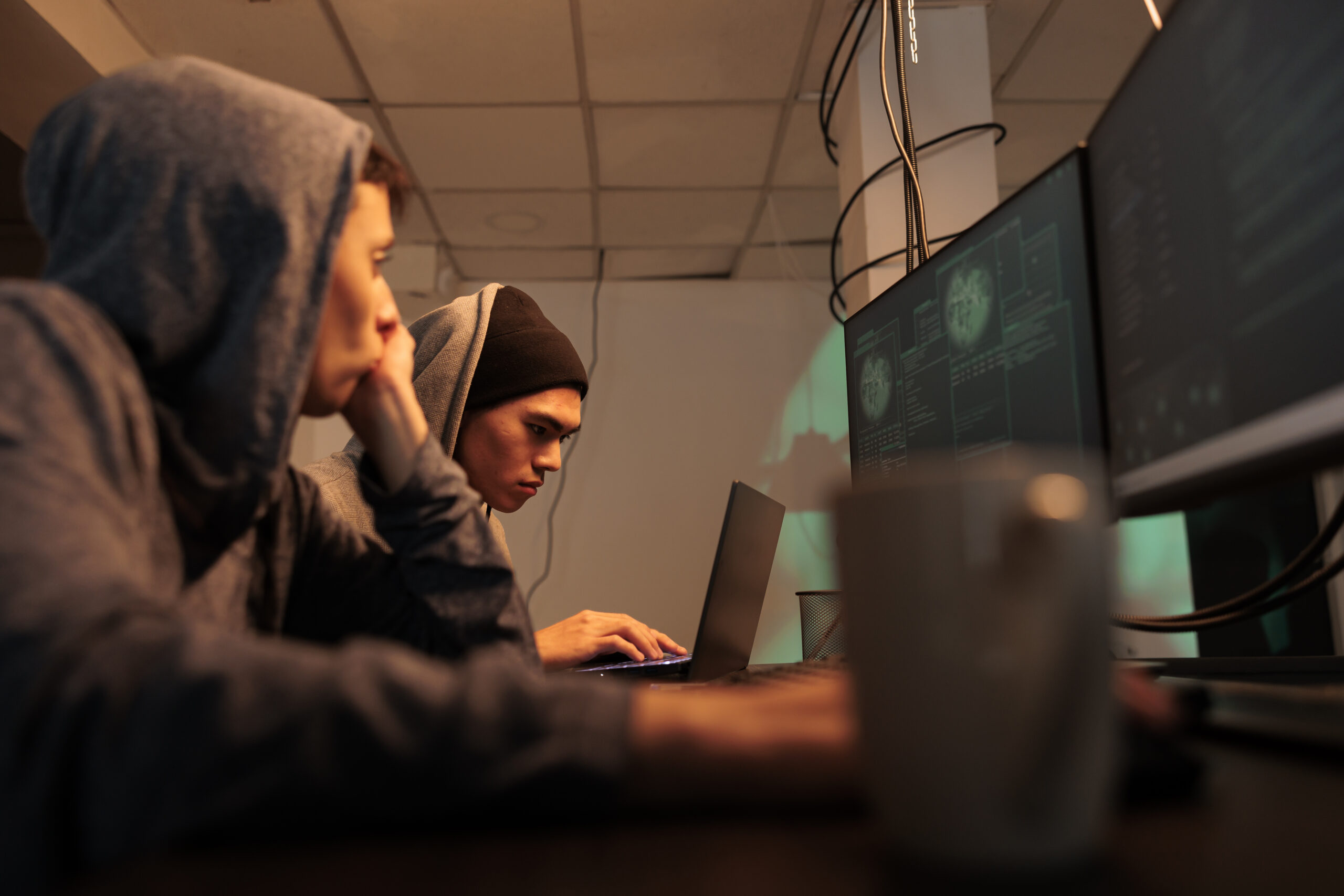 Jongeren met capuchons werken geconcentreerd achter laptops en meerdere schermen in een donkere ruimte