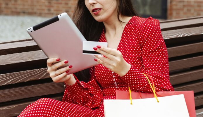 Vrouw zit op een bankje en doet een online aankoop met een tablet en creditcard, met een shoppingtas naast zich