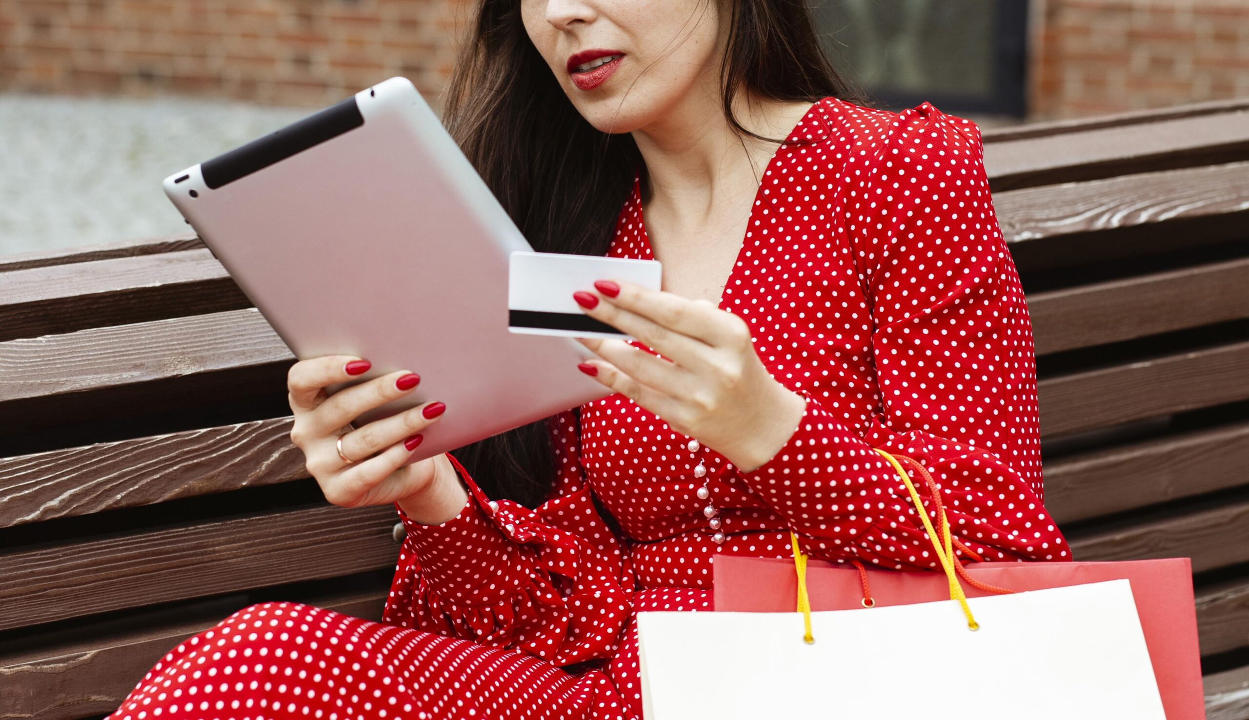 Vrouw zit op een bankje en doet een online aankoop met een tablet en creditcard, met een shoppingtas naast zich
