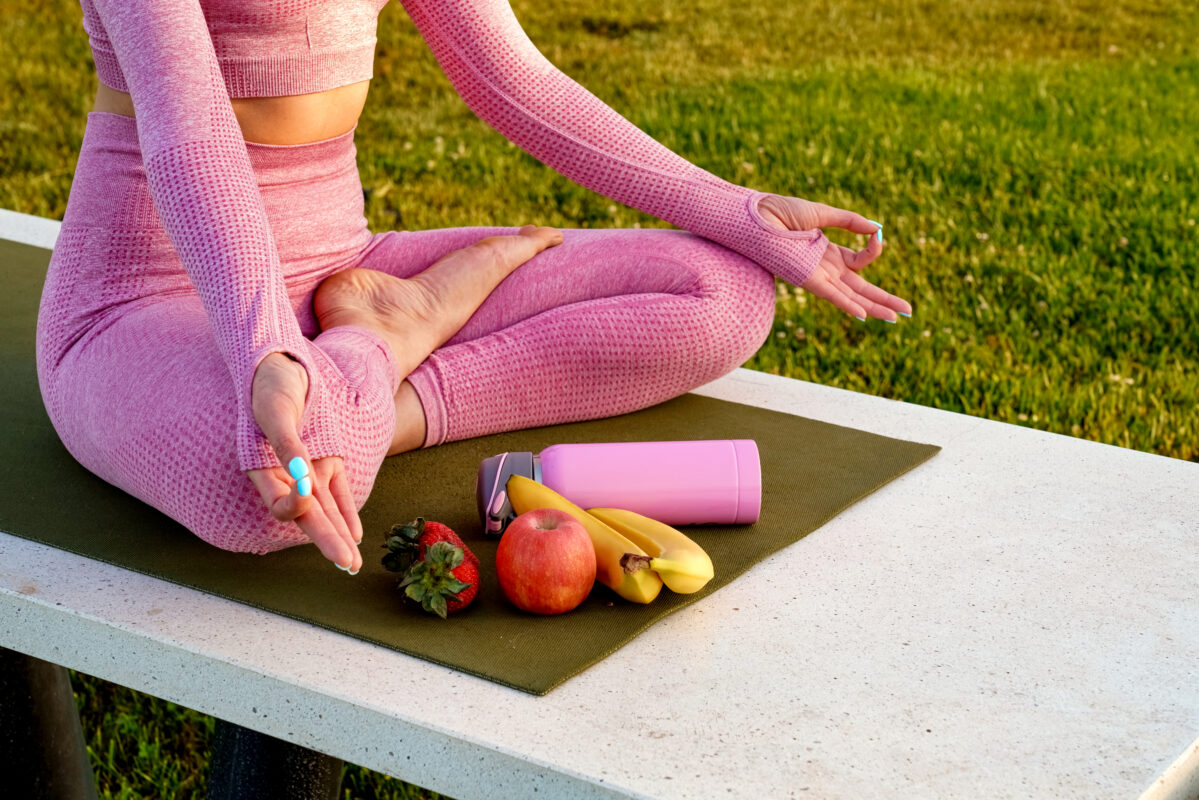 Jongere zit in yogahouding op een mat buiten met fruit, een waterfles en sportkleding als symbool voor een gezonde leefstijl.