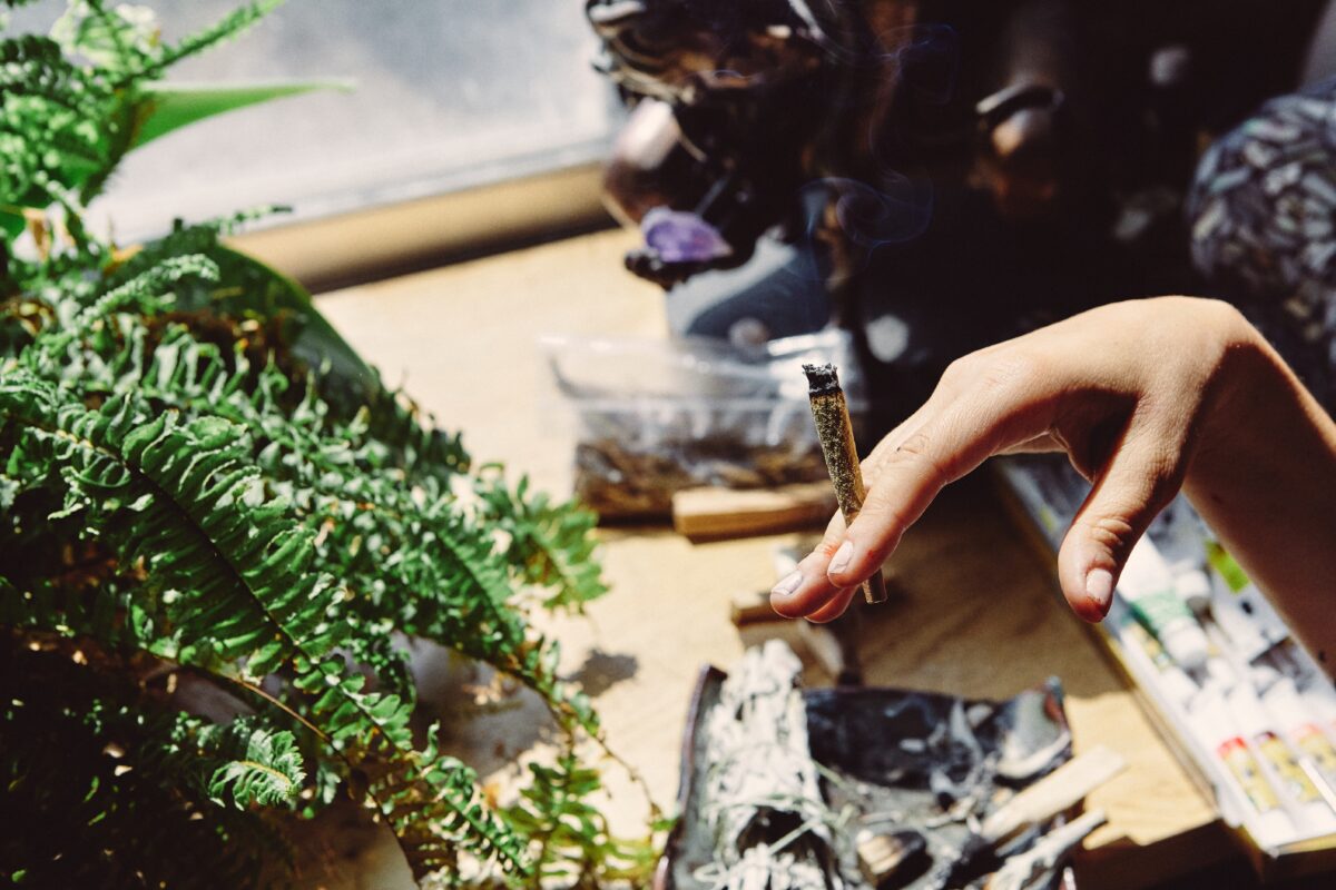 Hand met een opgerolde joint boven een tafel met kruiden en rook, als illustratie bij een artikel over synthetische cannabinoïden (spice) en drugsgebruik onder jongeren.