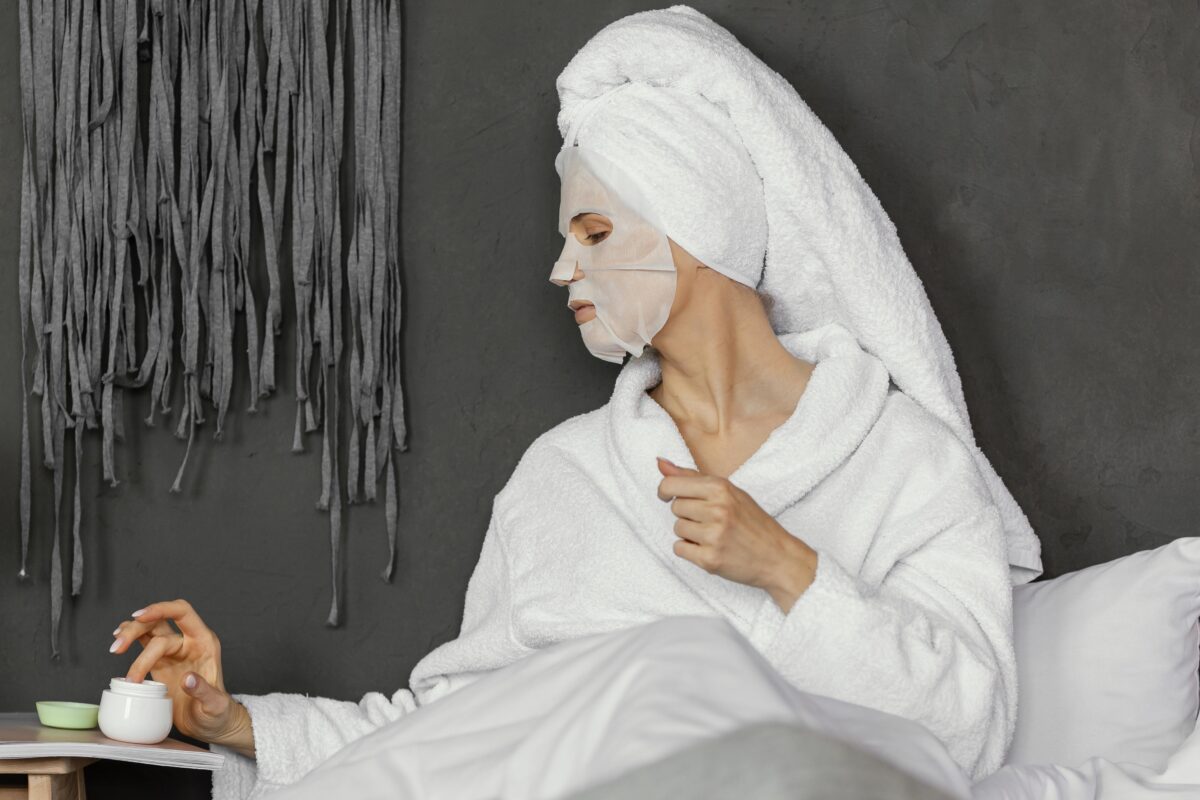 Persoon met een gezichtsmasker en handdoek om het haar brengt skincare aan in bed, passend bij de Morning Shed-trend op sociale media.