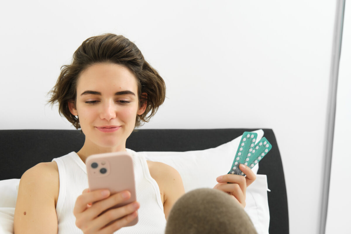 Jonge vrouw kijkt op haar smartphone terwijl zij een strip anticonceptiepillen vasthoudt. De afbeelding illustreert het zoeken naar gezondheidsinformatie of advies over anticonceptie via sociale media of internet.