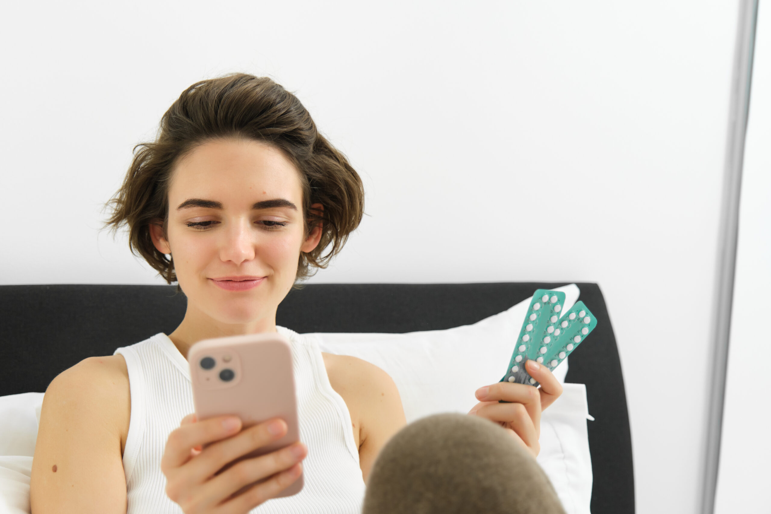 Jonge vrouw kijkt op haar smartphone terwijl zij een strip anticonceptiepillen vasthoudt. De afbeelding illustreert het zoeken naar gezondheidsinformatie of advies over anticonceptie via sociale media of internet.