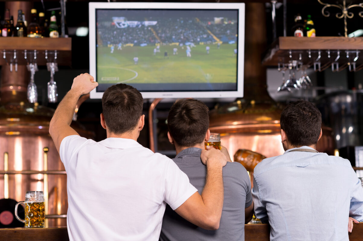 Drie personen kijken samen naar een voetbalwedstrijd op een groot scherm in een café of sportbar tijdens een internationaal voetbaltoernooi.