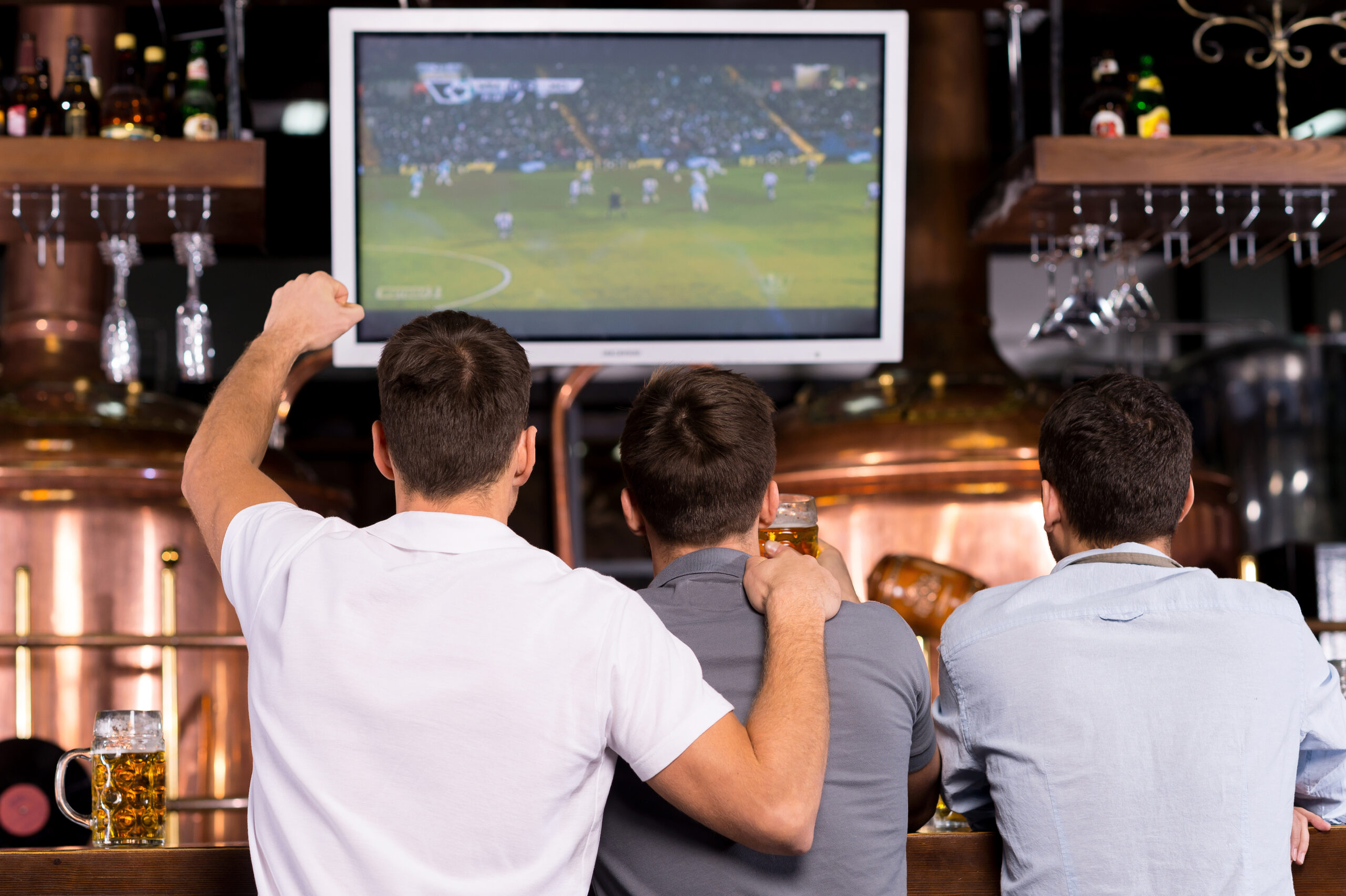 Drie personen kijken samen naar een voetbalwedstrijd op een groot scherm in een café of sportbar tijdens een internationaal voetbaltoernooi.