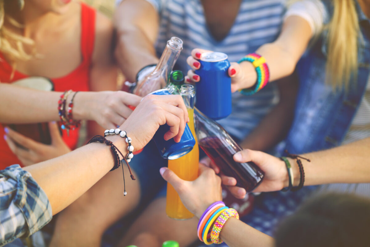 Groep jongeren proost met blikjes en glazen frisdrank en alcohol tijdens een feestje, als illustratie van alcoholgebruik onder jongeren.