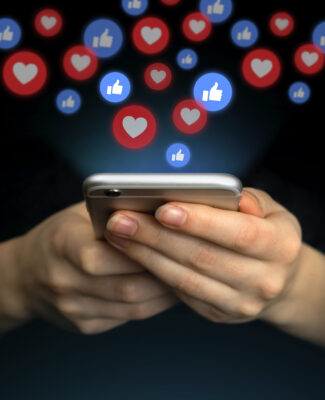 Handen houden een smartphone vast, terwijl likes en hartjes als sociale media-notificaties boven het scherm zweven. De afbeelding symboliseert de invloed van likes, views en online waardering.