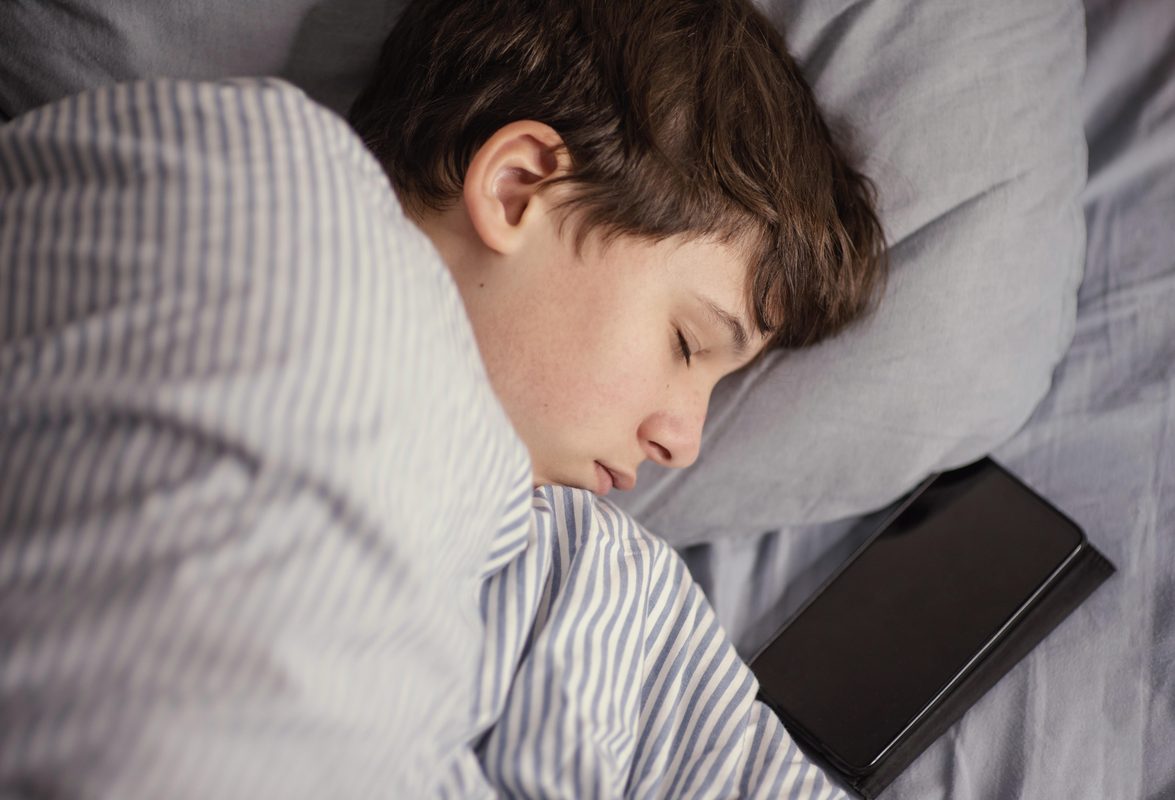 Slapende jongere met een smartphone naast het kussen, passend bij de sleepmaxxing-trend op sociale media.